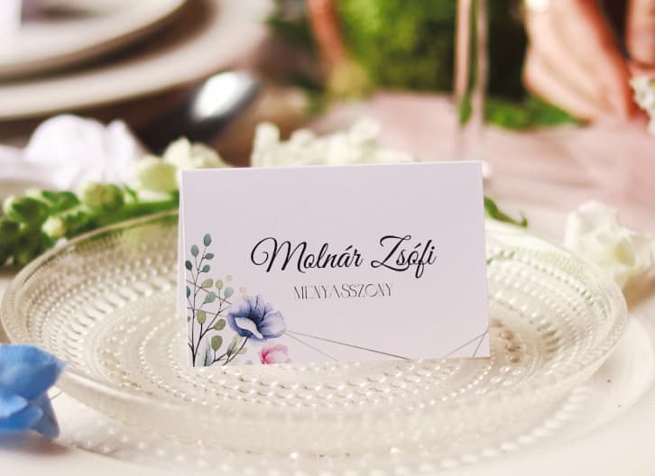 An elegant wedding placecard with floral designs on a transparent glass plate, surrounded by soft wedding decor.